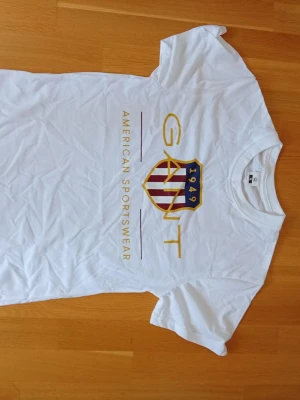 Vit GANT t-shirt med tryck - Snygg vit t-shirt från GANT med stort tryck framtill i guld, rött och blått. Trycket har texten 'GANT 1949 American Sportswear' och ett sköldmärke med ränder. Klassisk passform och rund hals. Perfekt till jeans eller shorts.