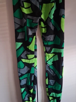 Färgglada leggings med grafiskt mönster - Säljer ett par leggings med coolt grafiskt mönster i svart, grönt, limegrönt och grått. Tight passform och stretchigt material som sitter skönt på benen. Perfekta för dig som vill sticka ut med din stil. Finns uppe på fler sidor så först till kvarn