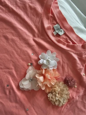 Croppad River Island t-shirt med 3D-blommor (Stl 40) ✨ - Söt rosa t-shirt med korta ärmar och flera 3D-blommor i olika färger och material på bröstet. T-shirten har en croppad passform och små pärldetaljer som ger extra wow-känsla. Perfekt för dig som vill sticka ut lite extra. Mått (liggande platt):Längd: 53 cm  Bredd (armhåla till armhåla): 51,5 cm  Längd från armhåla och ner: 26 cm