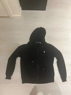 Svart hoodie från Polo Ralph Lauren - Säljer en svart hoodie från Polo Ralph Lauren med dragkedja och vit broderad logga på bröstet. Tröjan har huva med dragsko och ribbade muddar. Perfekt för en chill och stilren look.
