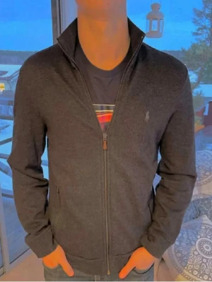 Mörkgrå ziptröja från Polo Ralph Lauren - Snygg mörkgrå ziptröja från Polo Ralph Lauren med hög krage och dragkedja framtill. Tröjan har två fickor med dragkedja och den klassiska broderade loggan på bröstet. Perfekt att slänga på sig över en t-shirt för en avslappnad look.