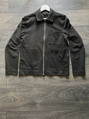 Svart overshirt  - Stilren svart overshirt från Elvine med rak passform, tre utanpåliggande fickor och dragkedja framtill. Tillverkad i ett mjukt tyg som känns bekvämt och passar perfekt till lager-på-lager. Klassisk krage och minimalistisk design.