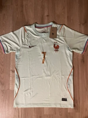 Frankrike fotbollströja Dembele #7 - Ljusblå matchtröja för Frankrikes landslag med Dembele och nummer 7 på ryggen. Tröjan har korta ärmar, tunna orange detaljer och broderad tupp-logga. Ribbade muddar med franska flaggans färger. Perfekt för fotbollsfans. HELT NY!