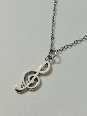 Silverfärgat halsband med nota - Snyggt halsband i silverfärg med ett hängsmycke format som en G-klav. Kedjan är supertunn men slitstark och smidig, och hänget har en stilren, blank finish. Perfekt för dig som älskar musik och vill ha en unik detalj till din outfit. Materialet rostfritt stål 