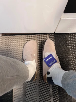 Beige Birkenstock tofflor i mocka - Säljer ett par snygga beigea Birkenstock tofflor i mjuk mocka. Modellen har en klassisk slip-in design med justerbart spänne och bekväm korksula. Perfekta för chill dagar hemma eller när du vill ha något enkelt men stilrent på fötterna.