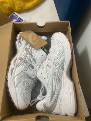 Vita ASICS Gel-Kayano 14 sneakers - Snygga vita ASICS Gel-Kayano 14 sneakers med silvriga detaljer och meshpaneler för extra ventilation. Skorna har en chunky sula i vitt och gummi, samt klassiska ASICS-stripes på sidorna. Perfekta för dig som gillar sportig och trendig stil.
