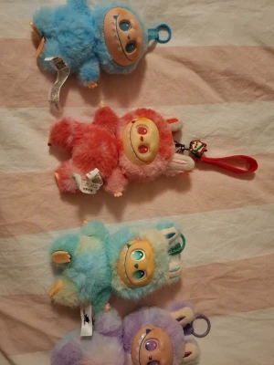 Färgglada fluffiga monster-nyckelringar -  ( den lila kan låta, sjunger då en labubu sång typ 30 sekunder.)  Fyra söta och fluffiga monster-nyckelringar i blått, rosa, turkos och lila. Varje figur har ett stort leende ansikte, mjuk päls och öron. Perfekt som accessoar till väskan eller nyckelknippan. Flera har färgade öglor för enkel upphängning.