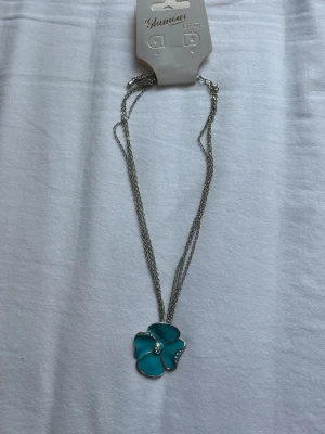 Silverhalsband med blå blomma - Snyggt halsband med en stor blå blomma som hänge. Blomman har silverfärgade kanter och små glittriga stenar i mitten. Kedjan är tunn och silverfärgad, vilket ger ett stilrent intryck. Perfekt för dig som vill ha en färgklick till din outfit.