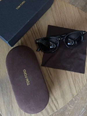 Tom Ford solglasögon - Snygga svarta solglasögon från Tom Ford med klassisk rundad form och mörka glas. Bågarna är i blank svart plast med silverdetaljer vid gångjärnen. Kommer med originalfodral och putsduk, perfekt för dig som vill ha en stilren accessoar.