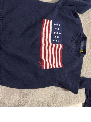 Marinblå stickad tröja Polo Ralph Lauren - Snygg marinblå stickad tröja från Polo Ralph Lauren med amerikansk flagga i rött, vitt och blått på bröstet. Tröjan har rund halsringning och ribbade muddar vid ärmslut och nederkant. Perfekt för en klassisk och avslappnad stil.