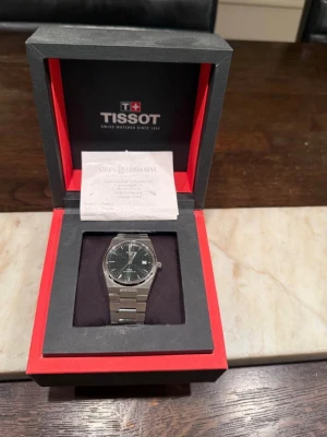 Tissot PRX silverfärgad klocka - Snygg Tissot PRX klocka i rostfritt stål med silverfärgad länk och svart urtavla. Klockan har en stilren och modern design med tydliga index och datumfönster. Perfekt accessoar för dig som gillar klassisk men trendig stil.