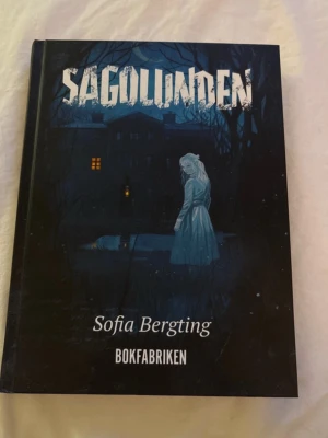 Sagolunden av Sofia Bergting - Sagolunden är en skräckroman av Sofia Bergting, utgiven av Bokfabriken. Omslaget är mörkt med blå och svarta toner och visar en mystisk flicka i vit klänning framför ett gammalt hus. Perfekt för dig som gillar spännande och kusliga berättelser.