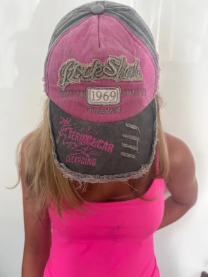 Grå och rosa vintagekeps - Säljer en grå och rosa keps med slitna detaljer och broderade texter framtill. Kepsen har en vintage look med fransiga kanter och tryck i vitt och rosa. Justerbar baktill och tillverkad i bomullstyg för en avslappnad streetstyle.