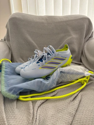 Vita Adidas Predator fotbollsskor - Snygga vita Adidas Predator fotbollsskor med gula och lila detaljer. Skorna har låg profil, snörning och en yttersula i gult för bra grepp på planen. Tillverkade i syntetmaterial och levereras med en matchande påse. Perfekta för dig som vill sticka ut på planen.