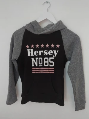 Svart och grå hoodie Hersey - Snygg hoodie från Hersey med svart kropp och grå ärmar och huva. Tryck framtill med rosa stjärnor, texten 'Hersey No85' och rosa ränder. Den är i storlek 12/13 years men funkar lika bra som XS.