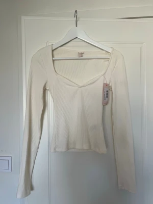 Vit ribbad långärmad topp - Trendig vit ribbad topp med lång ärm och snygg v-ringad halsringning. Figurnära passform och stretchigt material som ger en bekväm känsla. Perfekt att styla med jeans eller kjol för en clean look. Ny med prislapp kvar!
