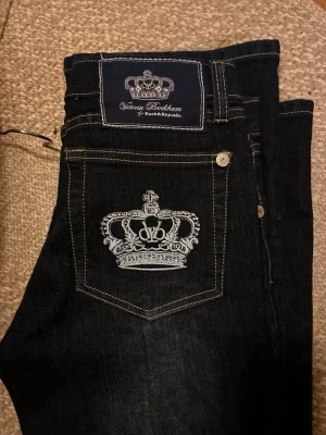 Mörkblå Victoria Beckham jeans - Snygga mörkblå jeans från Victoria Beckham för Rock & Republic. Jeansen har raka ben och coola broderade kronor på bakfickorna. Klassisk femficksmodell med kontrastsömmar och detaljer i silver. Perfekta för dig som vill sticka ut lite extra.