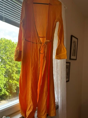 Orange långklänning med knytband - Långklänning i stark orange färg med krage, långa ärmar och knytband i midjan. Klänningen har en lätt skrynklig struktur och knappar vid ärmsluten. Perfekt för sommarens varma dagar.
