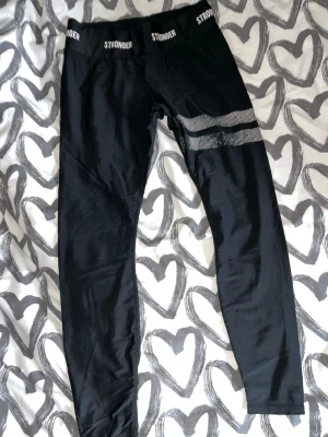 Leggings  - Snygga svarta leggings från Stronger med vita prickiga ränder på ena benet och logga i midjan. Stretchigt material som sitter tight och bekvämt. Perfekta för träning eller chill. Midjehög modell med elastisk linning. Använt skick men inget trasigt.