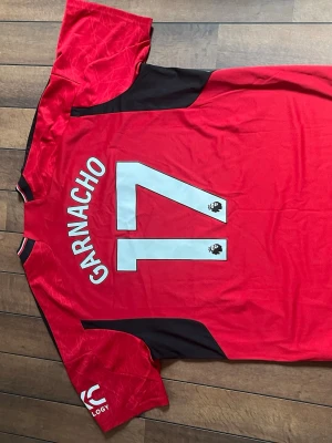 Manchester United matchtröja Garnacho - Säljer en röd Manchester United matchtröja från Adidas med Garnacho och nummer 17 på ryggen. Tröjan har svarta detaljer, klubbmärke på bröstet och TeamViewer som sponsor. Perfekt för dig som älskar fotboll och vill stötta ditt lag på matchdag.