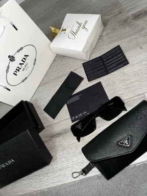 Svarta solglasögon från Prada - Snygga svarta solglasögon från Prada med klassisk rektangulär form. Glasögonen har diskret Prada-logga på skalmen och kommer med ett svart fodral i strukturerat material samt originalförpackning och tillbehör.