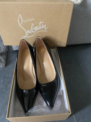 Svarta lackpumps från Louboutin - Snygga svarta pumps i lack från Christian Louboutin med klassisk spetsig tå och låg klack. Skorna har den ikoniska röda sulan och elegant siluett, perfekta för att lyfta vilken outfit som helst.