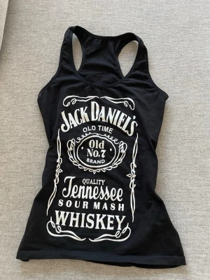 Jack Daniel's svart linnetopp - Svart linnetopp med Jack Daniel's-tryck i vitt framtill och mindre logga bak.   Har en slim passform. Perfekt för dig som gillar statement-plagg och rockig stil.