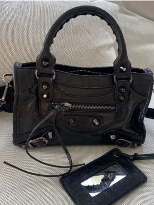 Svart handväska med detaljer - Cool svart handväska i läderlook med silvriga metalldetaljer, dragkedja framtill och matchande avtagbar axelrem. Väskan har två handtag och en tillhörande liten plånbok med spegel. Perfekt för dig som gillar edgy och trendig stil.