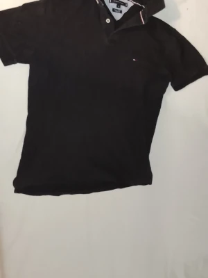 Svart pikétröja Tommy Hilfiger - Svart pikétröja från Tommy Hilfiger med klassisk krage och knappar framtill. Tröjan har diskret broderad logga på bröstet och rödvit detalj på kragen. Tillverkad i bomull och har korta ärmar för en clean och tidlös stil.