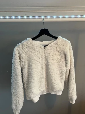 Vit fluffig teddyjacka med dragkedja - Supermjuk och fluffig vit teddyjacka med dragkedja framtill. Jackan har rund halsringning, långa ärmar och ribbade muddar vid ärmslut och nederkant. Perfekt för dig som vill ha en cozy och trendig look.