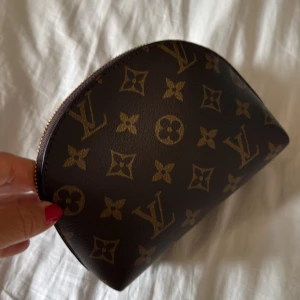 Ny brun liten necessär el sminkväska med monogram mönster i trendig modell  - Säljer en klassisk brun necessär med ikoniskt monogrammönster i guld. Halvmåneformad modell med dragkedja upptill. Den är tidlös och ger en exklusiv känsla. Perfekt för smink eller småprylar.