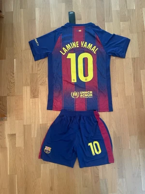 Lamine Yamal FC Barcelona hemmaställ - Säljer ett FC Barcelona matchställ med Lamine Yamal och nummer 10 på ryggen. Setet består av en kortärmad tröja och shorts i blått och vinrött med gula detaljer. Tröjan har klubbmärke och sponsorloggor, och shortsen har klubbmärke och nummer 10.