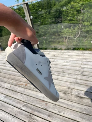 Golden goose skor - Snygga vita sneakers med grå stjärndetalj på sidan och grå häl. Skorna har grå skosnören och svart insida. Yttersulan är beige med svart undersida. Perfekta för en trendig och avslappnad stil.