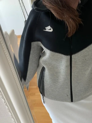 Nike hoodie i svart och grå - Nike hoodie med dragkedja i svart och ljusgrå färgblock. Stor vit Nike-logga på bröstet, huva med snörning och två fickor framtill. OBS! Fläck på ena sidan. Jag köpte den för 1 500 