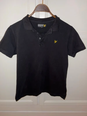 Svart pikéskjorta Lyle & Scott - Svart pikéskjorta från Lyle & Scott med klassisk krage, korta ärmar och knappar framtill. stilren och enkel design, passar till dig med storlek 164-176.