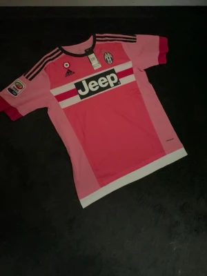 Juventus rosa matchtröja Pogba #10 - Juventus rosa matchtröja från Adidas med Pogba och nummer 10 på ryggen. Tröjan har svarta och vita detaljer, Jeep-logga på bröstet och Serie A-märke på ärmen. Tillverkad i lätt funktionsmaterial, perfekt för fotbollsträning eller match.