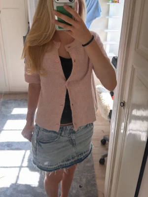 Ljusrosa ribbad kofta med knappar - Supersöt ljusrosa kofta med ribbad struktur och rund hals. Koftan har korta ärmar och stängs med matchande rosa knappar framtill. Perfekt att slänga över ett linne eller t-shirt för en chill look.