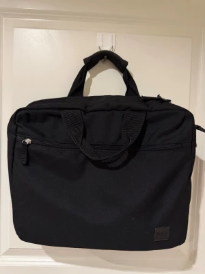 Stor svart datorväska från Hugo Boss - Stilren svart datorväska från Hugo Boss i slitstarkt tygmaterial. Väskan har dubbla handtag, en ytterficka med dragkedja och diskret logotyp framtill. Perfekt för att bära laptop och tillbehör till skolan eller jobbet. Får plats med ganska mycket.