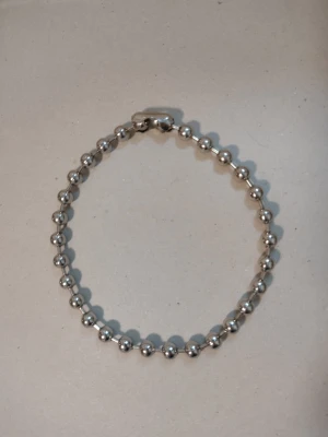 Grov silverfärgad kedja/kulkedja i metall - silverhalsband/pärlhalsband - Halsband/halskedja i glansig metall med runda, silverfärgade "pärlor"/kulor. Klassisk kulkedja i en grövre modell, med ett smidigt lås. Känner tyvärr inte till vad det är för typ av metall. Ca 43,5 cm lång när kedjan är knäppt och väger ca 70 gram. Kulorna är ca 1 cm i diameter. Sista kulan - den som används för att låsa kedjan - är lite sliten/repig.