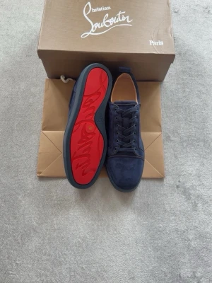 Mörkblå Christian Louboutin sneakers - Snygga mörkblå sneakers från Christian Louboutin med klassisk röd sula och diskret logga på sidan. Skorna har en låg profil, rund tå och är tillverkade i mocka och läder. Perfekta för dig som vill sticka ut med stilrena detaljer.