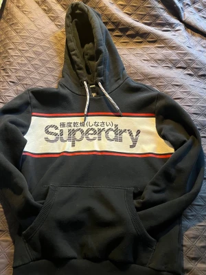 Mörkblå Superdry hoodie med tryck - Mörkblå hoodie från Superdry med stor logga och japansk text på bröstet. Har vit panel med röda linjer, justerbar huva med snören och en klassisk magficka. Perfekt för en avslappnad streetwear-look. Mjuk bomullskänsla och ribbade muddar.