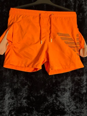 Oranga badbyxor med svart logga - Snygga oranga badbyxor med svart randigt logotryck på vänster ben. De har elastisk midja med dragsko och sidofickor. Materialet är lätt och snabbtorkande, perfekt för stranden eller poolen. Skickas snabbt inom 1-2 dagar. Djur- och  