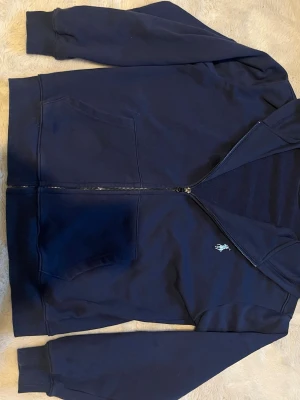 Marinblå hoodie från Polo Ralph Lauren - Snygg marinblå hoodie från Polo Ralph Lauren i storlek S. Dragkedja framtill, två stora fickor och klassisk huva. Liten broderad logga på bröstet. Tillverkad i mjuk bomull för skön känsla och avslappnad stil. Ganska liten i storleken har 1 liten fläck fram på bröstet typ.
