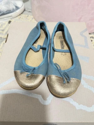 Blå ballerinaskor med guldtå - Snygga ballerinaskor i ljusblått tyg med metallic guld på tån och en liten rosett framtill. Skorna har en klassisk rundad form och elastiskt band över foten för extra komfort. Perfekta för dig som vill ha en stilren men ändå unik look.