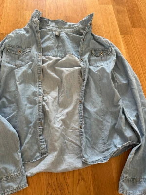 Ljusblå jeansskjorta med knappar - Snygg ljusblå jeansskjorta med långa ärmar och två bröstfickor med knappstängning. Skjortan har klassisk krage och knappar framtill. Perfekt att styla lager på lager för en avslappnad look.