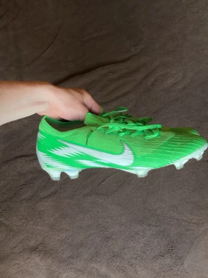 Nike vapor ”Naija” - Snygga gröna fotbollsskor från Nike med vit sula och vita detaljer på sidan. Skorna har snörning och är designade för spel på gräs. Ovandelen är i syntetmaterial med en modern, aerodynamisk form och tydliga mönster för extra grepp på bollen.