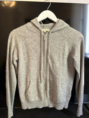 Grå hoodie i kashmir från Cozy Cashmere - Mysig grå hoodie från Cozy Cashmere i mjuk kashmir. Hoodien har dragkedja framtill, två fickor och långa ärmar. Perfekt för dig som gillar en clean och chill stil med premiumkänsla. Snygg att bära till jeans eller mjukis.