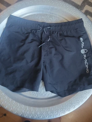 Svarta badbyxor Sail Racing - Snygga svarta badbyxor från Sail Racing med vitt logotryck på ena benet och matchande dragsko i midjan. Klassisk modell med elastisk midja och kortare ben. Perfekta för stranden eller poolen. Andvända 2 g.