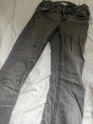 Grå raka jeans med fem fickor - Säljer ett par grå jeans med klassisk femficksdesign och raka ben. Jeansen har silverfärgade knappar och detaljerade sömmar vid fickorna. Perfekta för en avslappnad och stilren look.