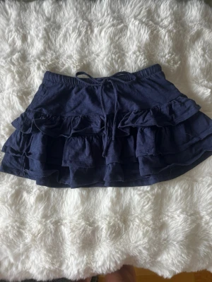Mörkblå volangkjol från Gina Tricot - Supersöt mörkblå kortkjol från Gina Tricot med flera lager volanger och snörning i midjan. Använd max 2 gånger.
